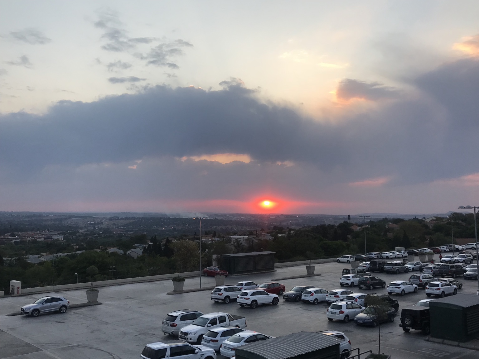A bright red evening sun beneath broad clouds over a city and parked cars.
